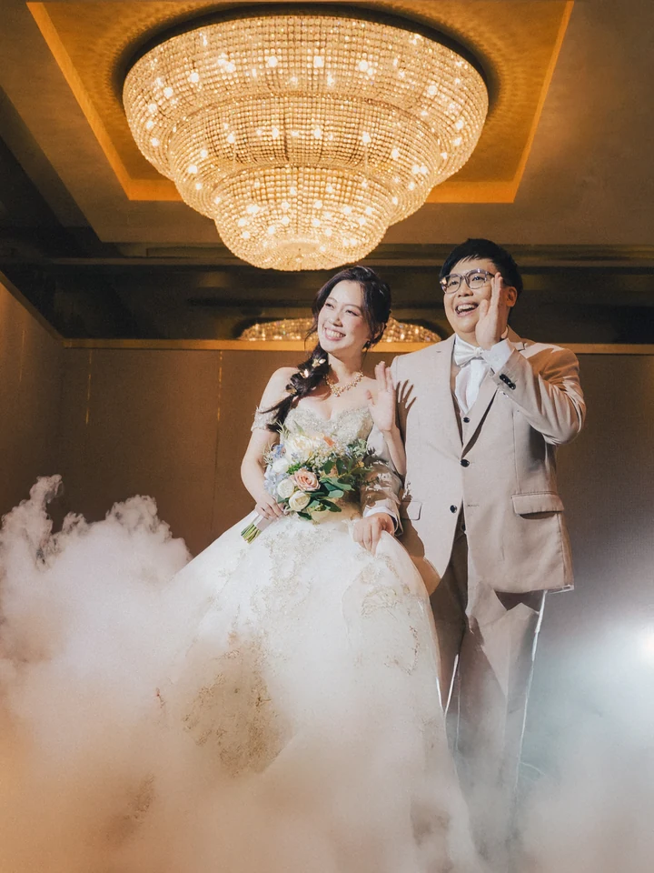 A wedding couple waving beneath a chandelier as mist fills the ballroom